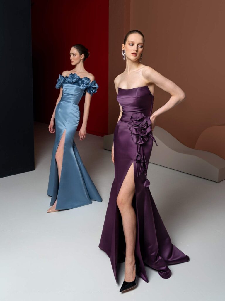 Two women in luxurious evening gowns, one in a deep purple with a thigh-high slit and floral accents, and the other in a soft blue with off-the-shoulder ruffles, showcasing different styles for Engagement Party Dress Codes.