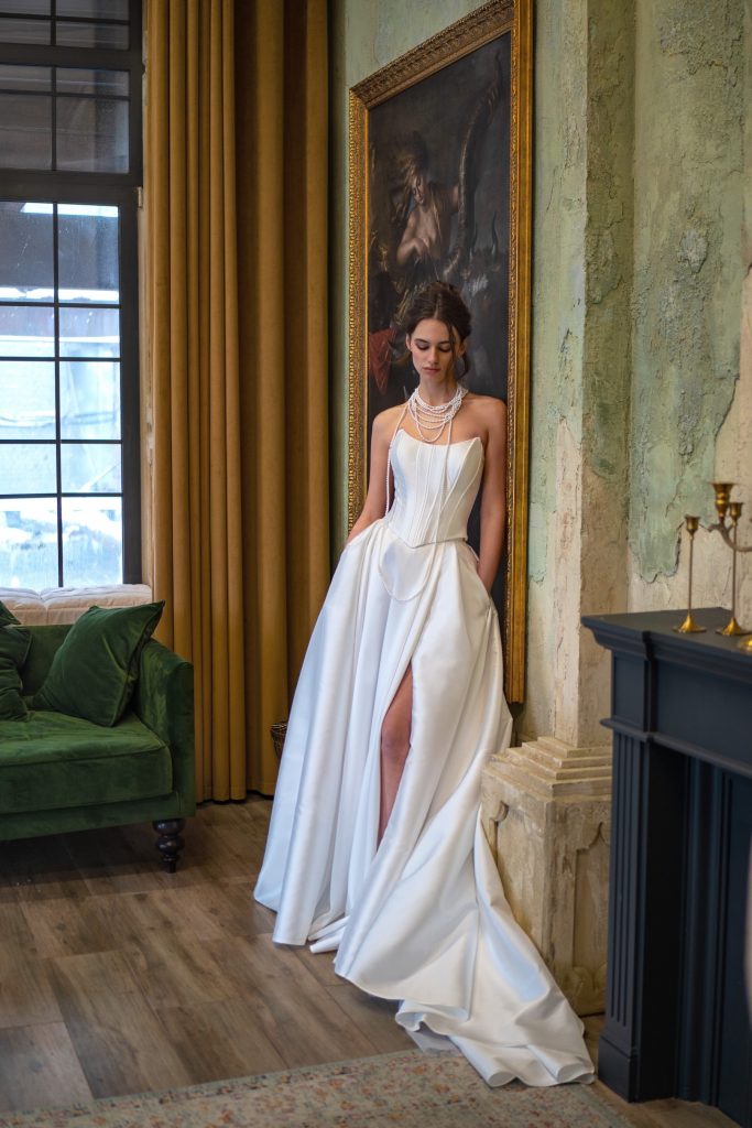 A luxurious satin wedding gown with a corset bodice and high slit, paired with pearl necklaces for a timeless bridal style. Inspired by Selena Gomez Wedding Dress, photographed against a regal interior with a classic portrait and golden drapery.