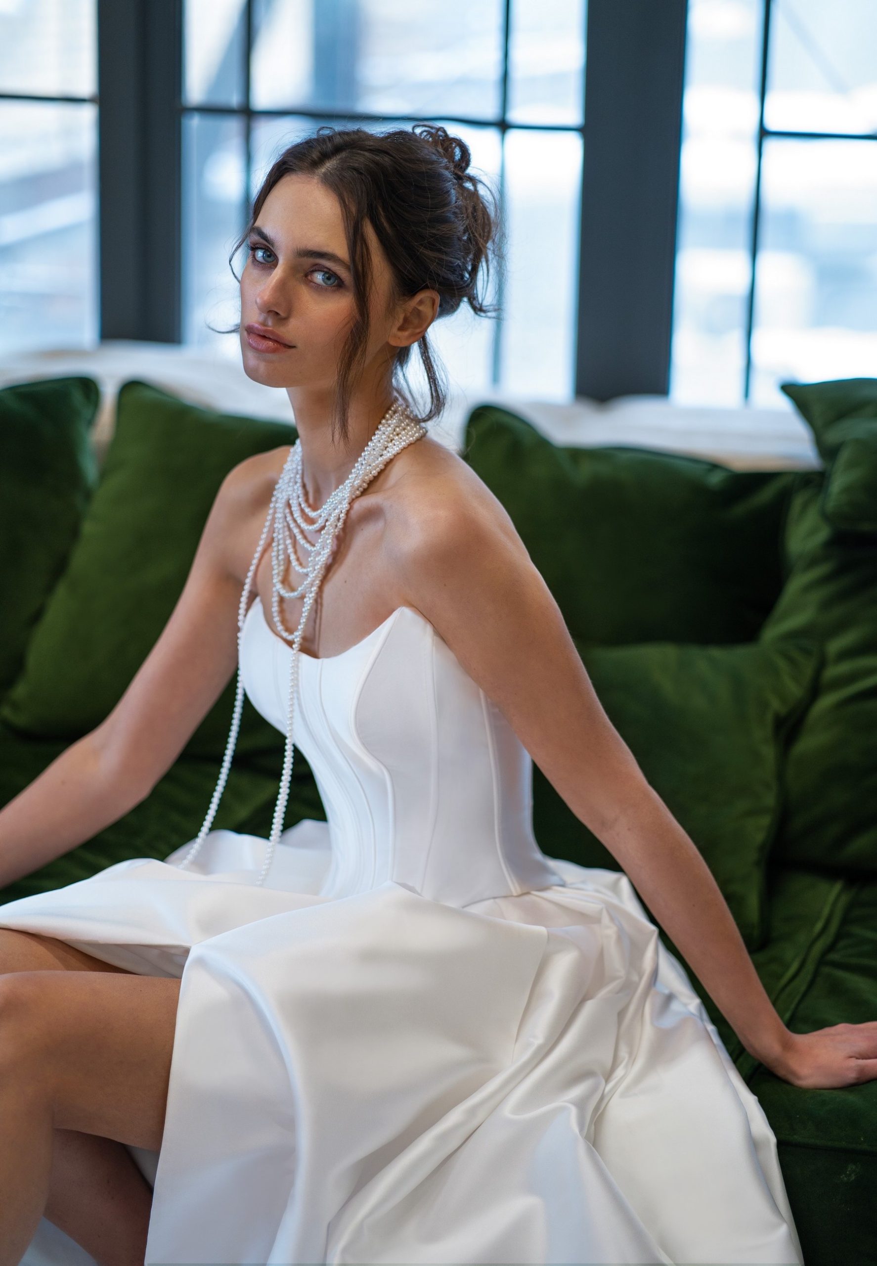 A chic strapless satin wedding dress featuring a fitted corset bodice and layered pearl necklaces. A modern take on bridal elegance reminiscent of Selena Gomez Wedding Dress. Captured in a cozy, green velvet couch setting.