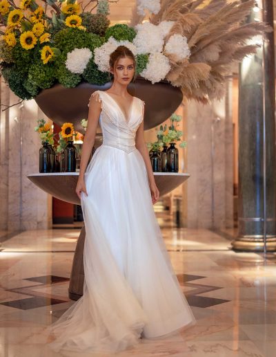 Bride in a simple wedding dress adorned with pearls, standing under a floral arrangement at Papilio Boutique.