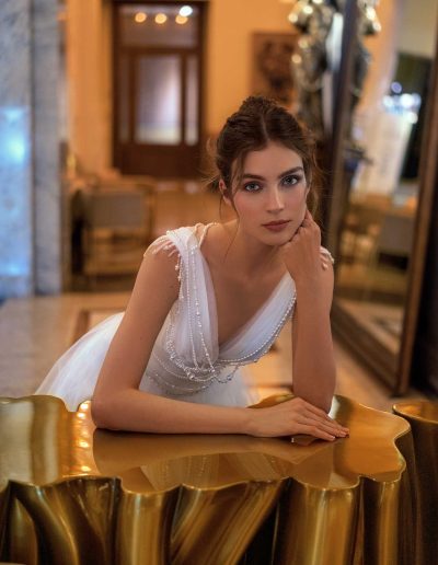 Model wearing a simple wedding dress with pearl decor, leaning on a golden table at Papilio Boutique Toronto bridal store.