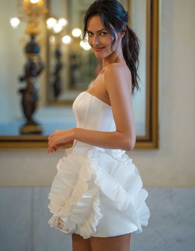 A captivating bride in a stylish mini satin wedding dress adorned with ruffles and a large floral accent on the hip, looking over her shoulder with a playful smile, in a luxurious hallway with reflective mirrors.