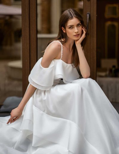 Elegant bride standing in organza gown with off-the-shoulder detail, by Papilio Boutique Toronto bridal store, in a serene outdoor setting.